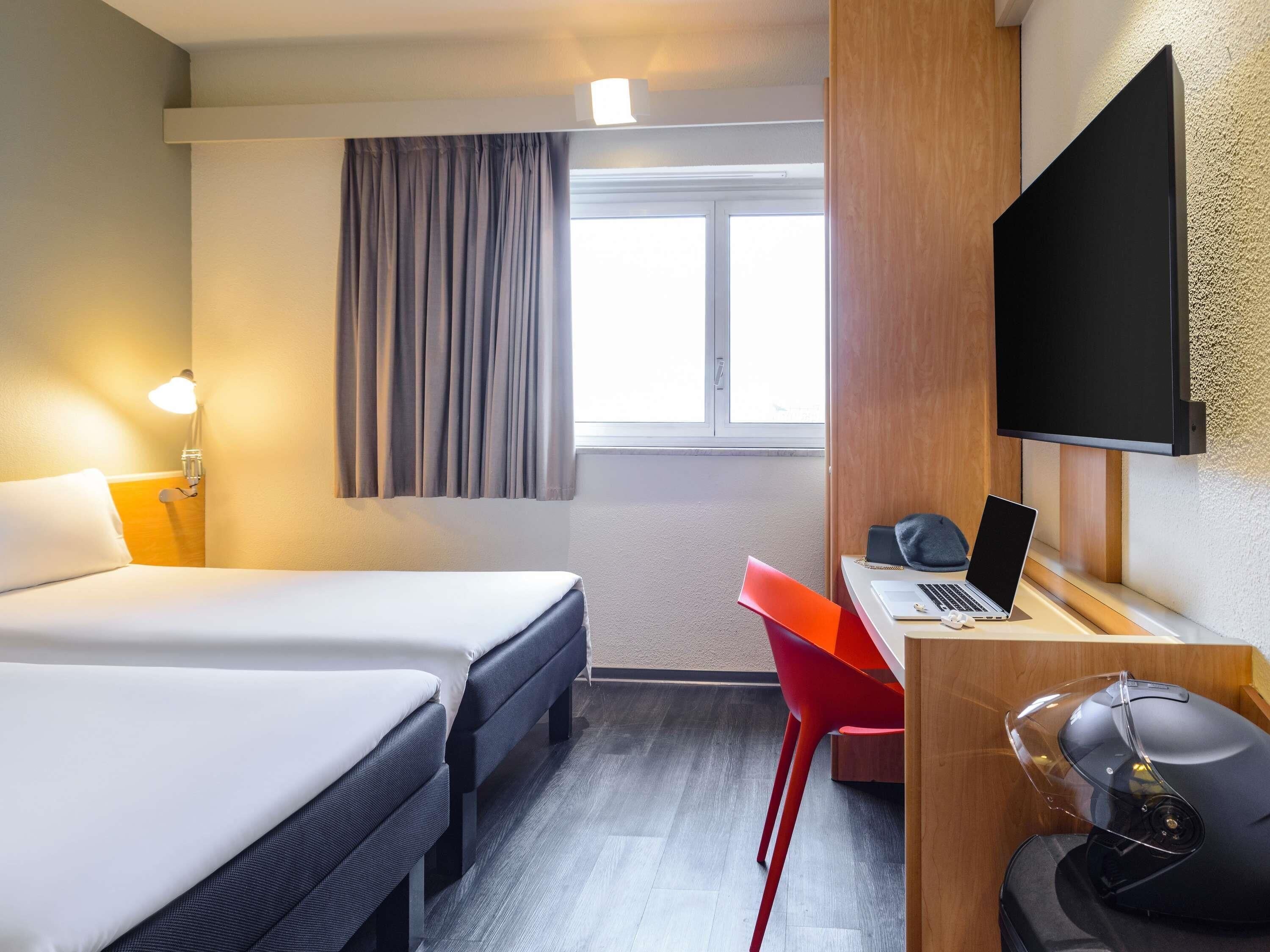 A hotel room with two beds, a desk with a laptop, a TV, and a red chair.
