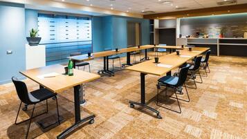 Un salón de conferencias con varias mesas y sillas, un proyector y una pared con ventana y persianas.