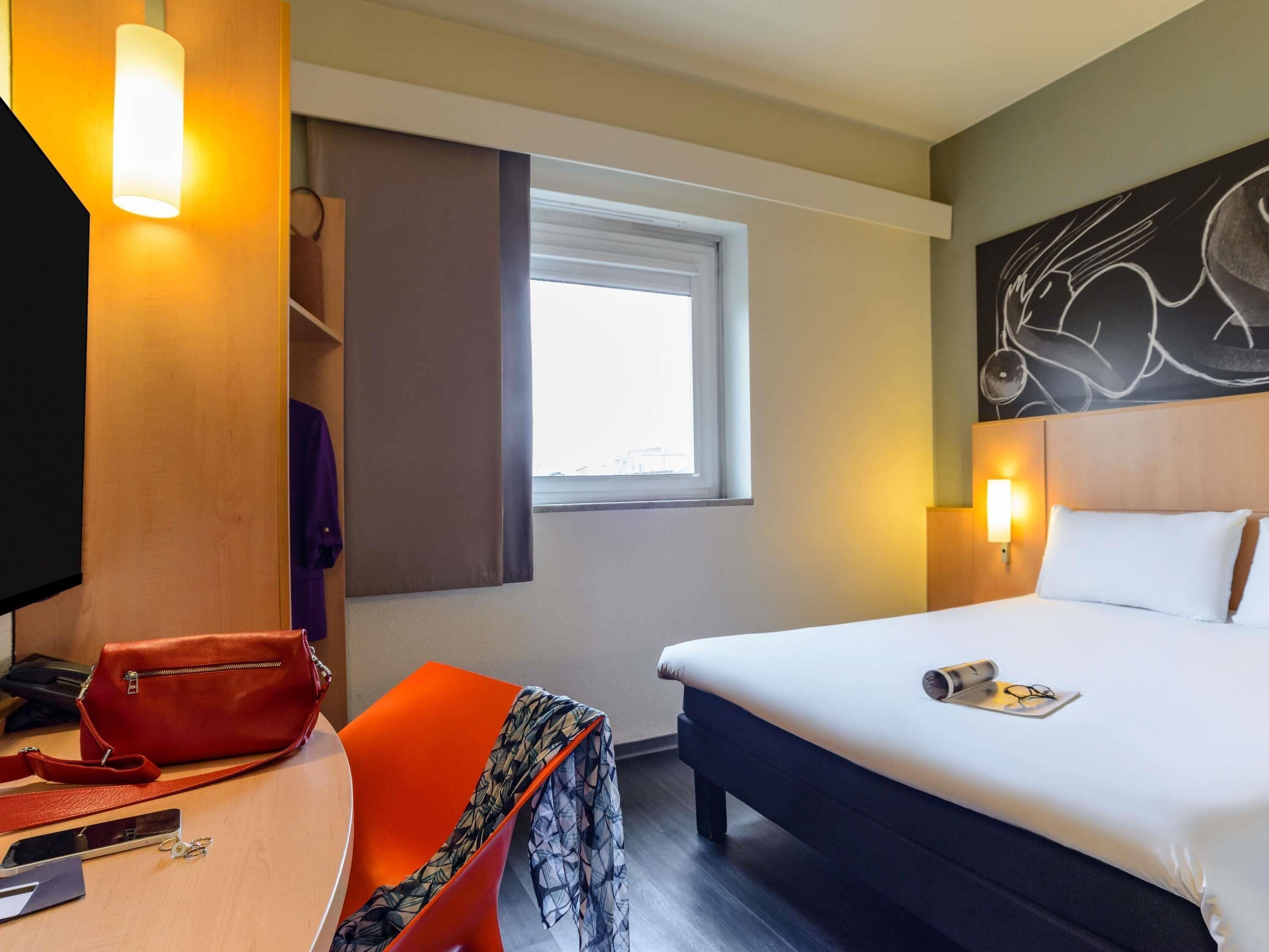 Una habitación de hotel con una cama, un escritorio con una silla, un perchero y una ventana.