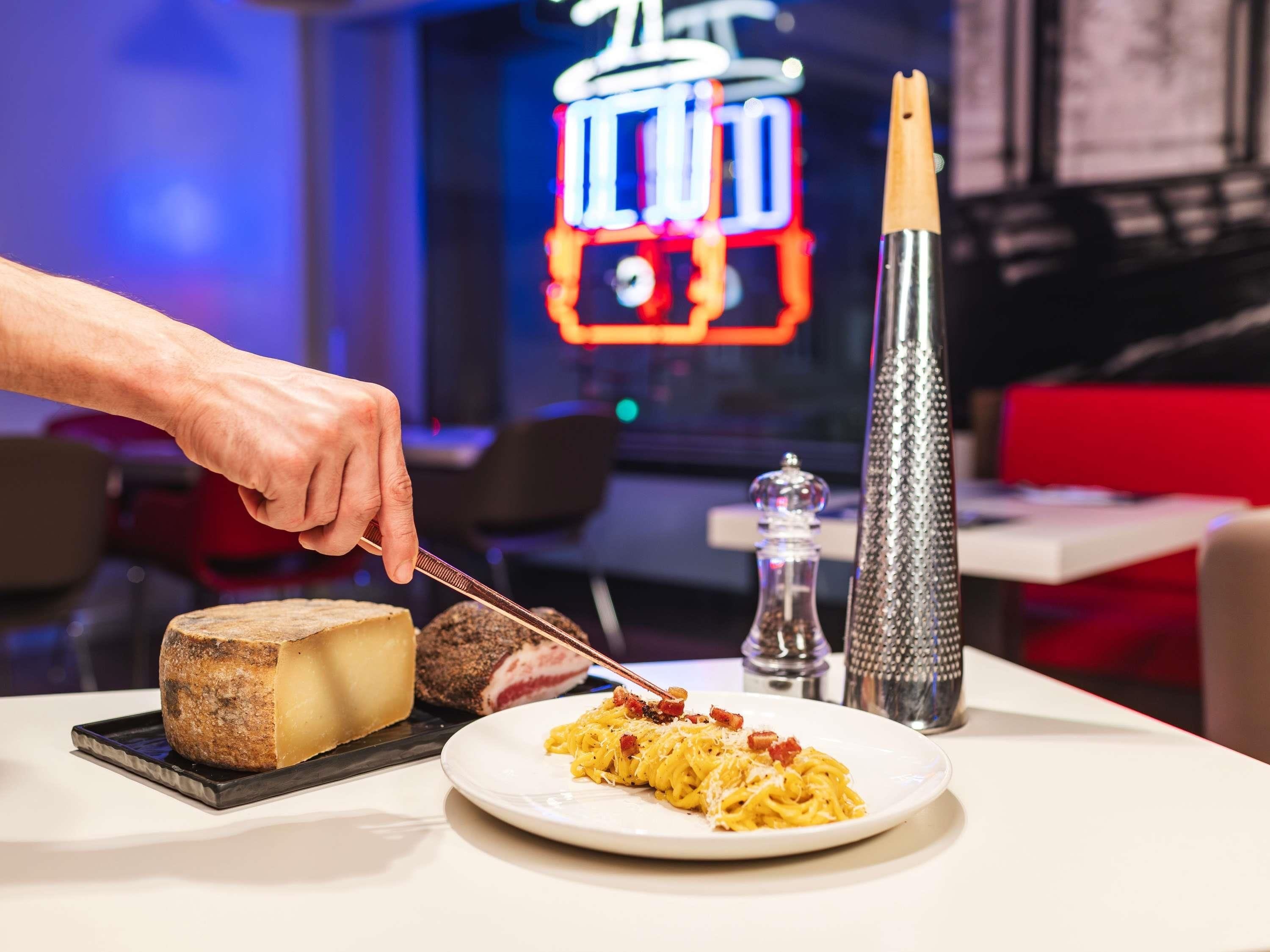 A plate of spaghetti with a knife cutting it, a cheese wheel, and a salt shaker on a table.