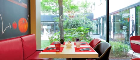 A restaurant with red seating, a table with a red placemat, and a view of a garden and buildings outside.