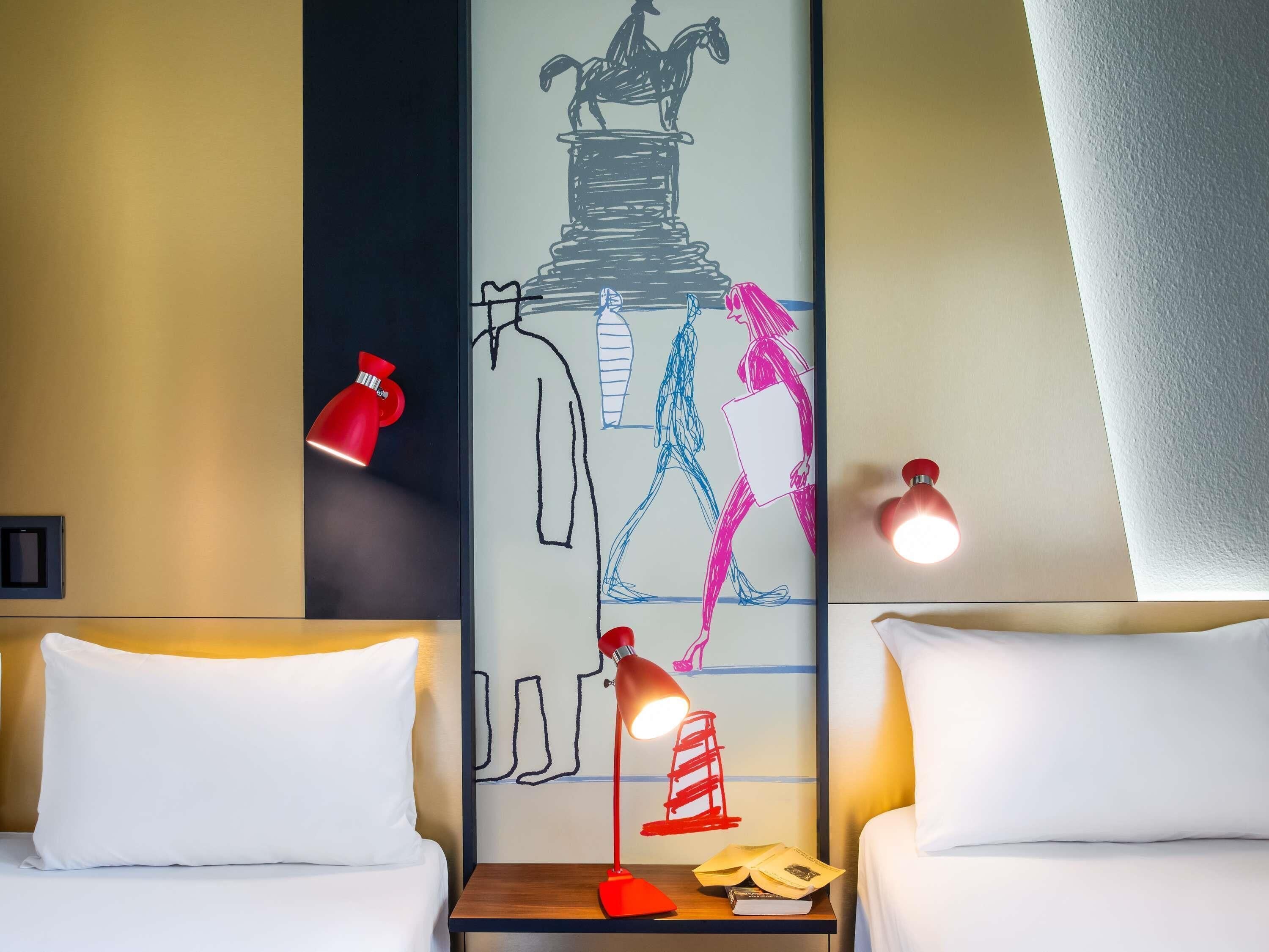 Habitación de hotel con dos camas, una mesita de noche con lámpara, una pared con mural y dos lámparas de pared.