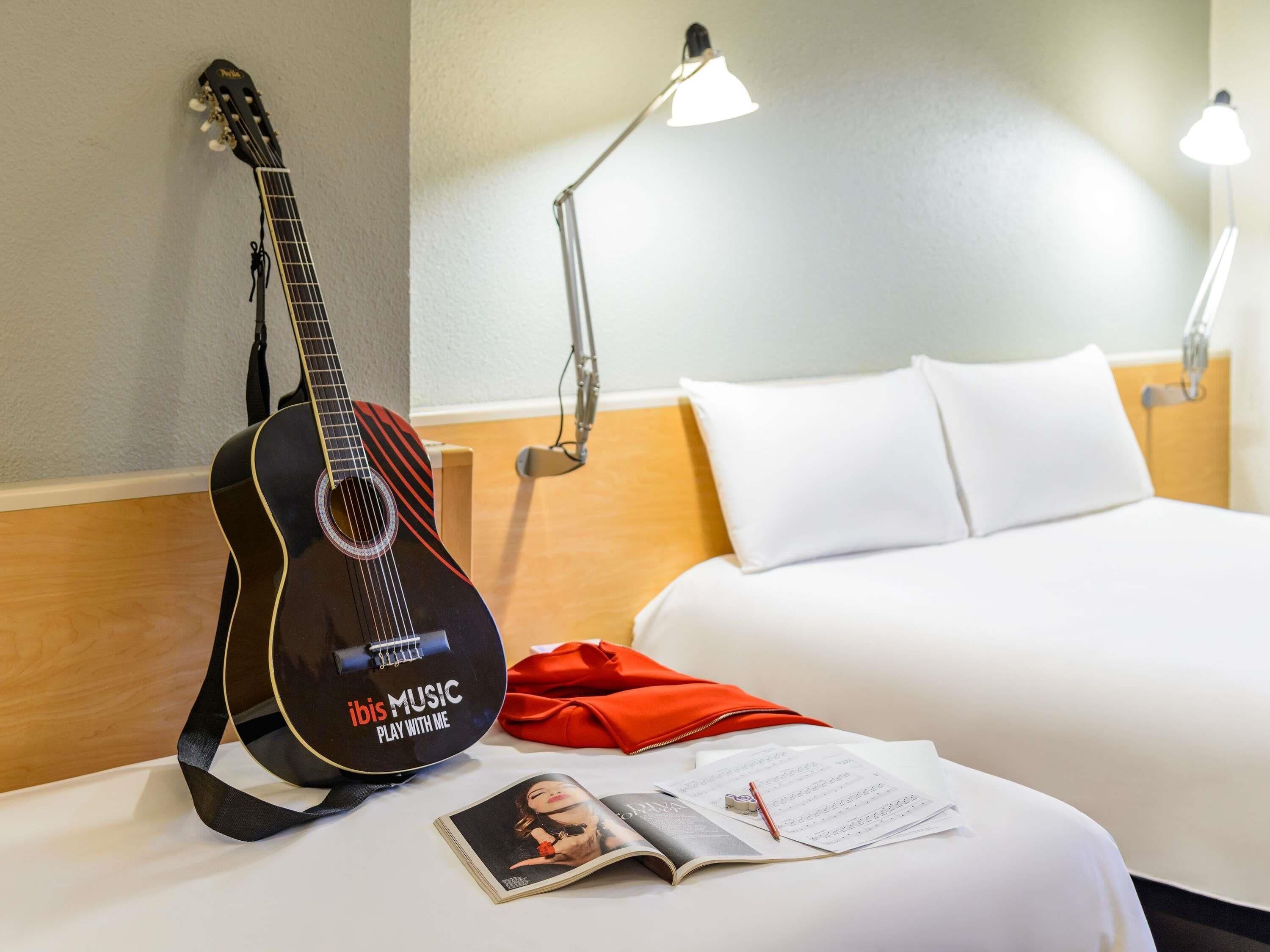 Een hotelkamer met een bed, een gitaar, een lamp, een rode tas en een tijdschrift.