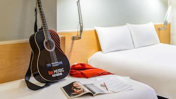 A hotel room with a bed, a guitar, a lamp, a red bag, and a magazine.