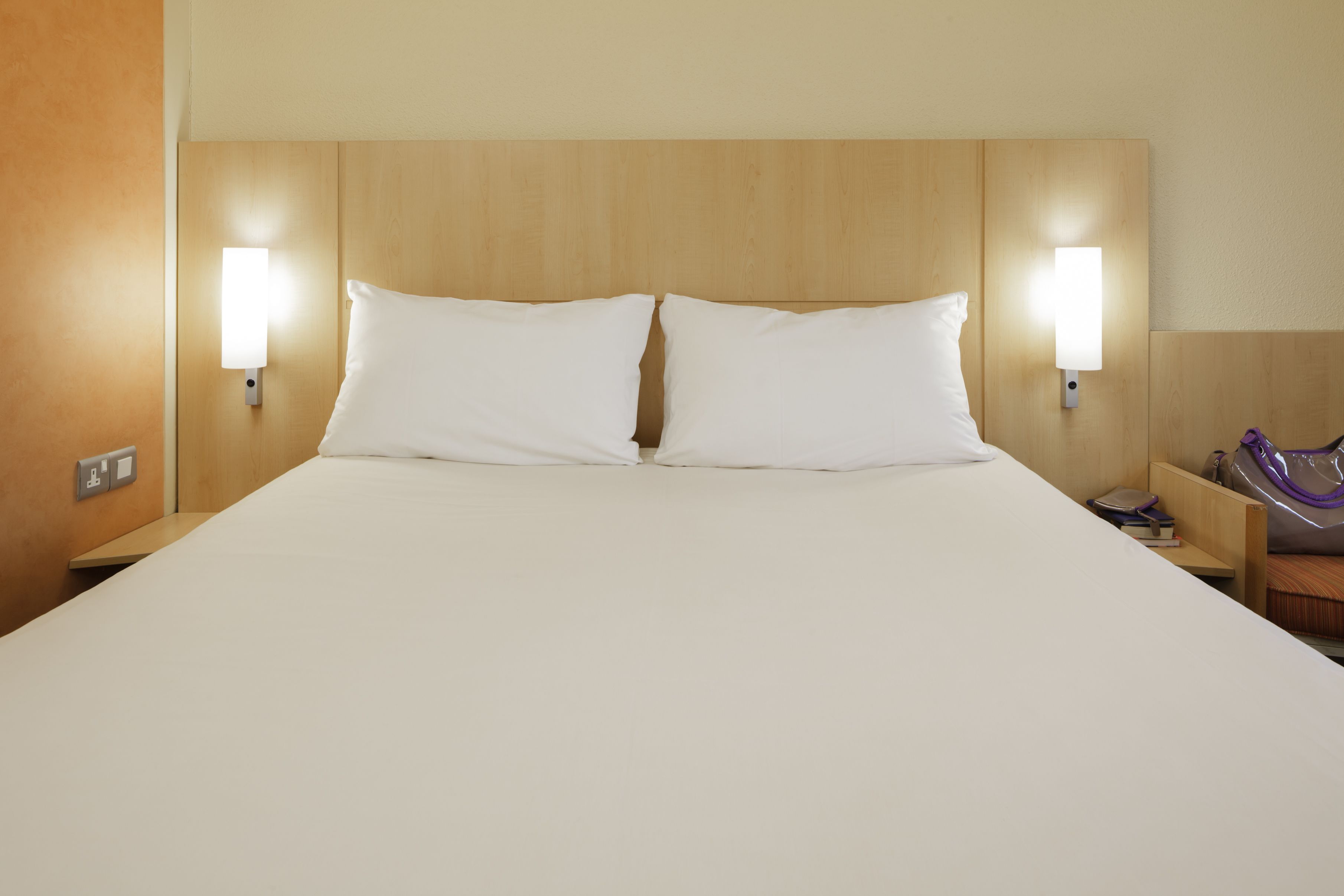 A hotel room with a large bed, two bedside lamps, a wooden headboard, and a nightstand with a lamp and a purple bag.