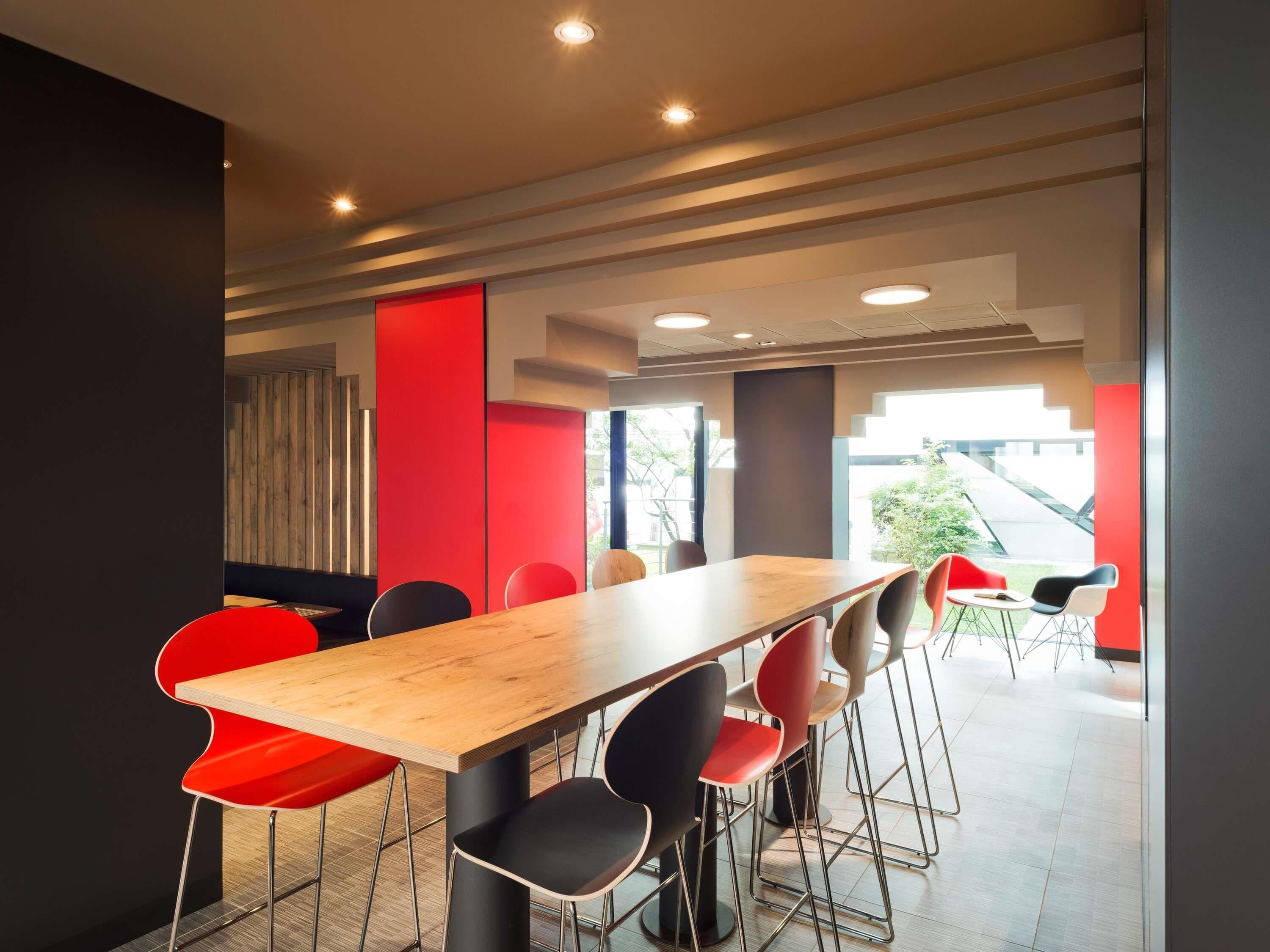 A modern conference room with a long wooden table, red and black chairs, and a view of the outdoors through large windows.