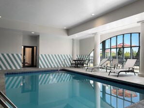 An indoor swimming pool with a tiled border, lounge chairs, and a glass wall with a view.