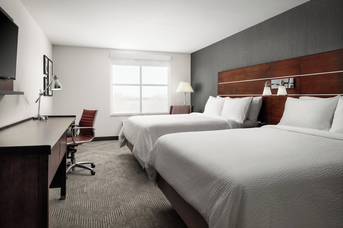 A hotel room with two beds, a desk, a chair, a lamp, and a window.