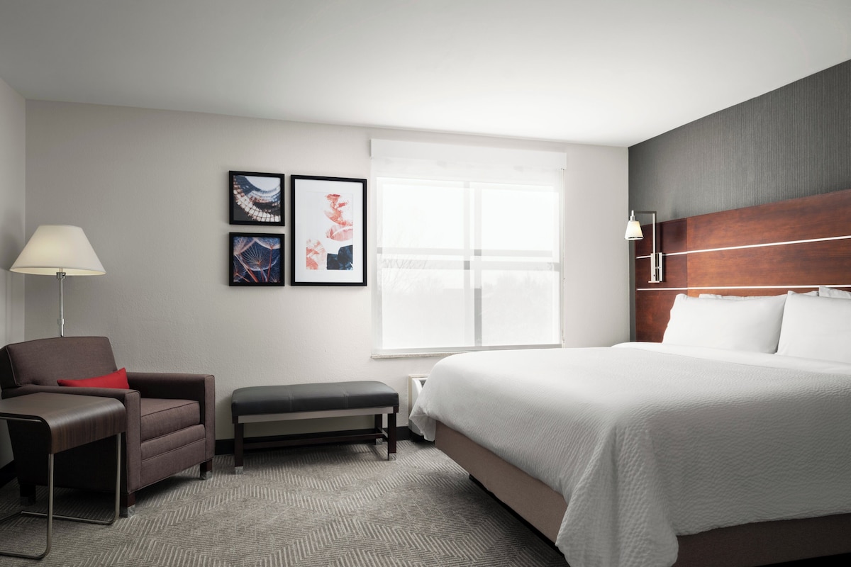 A hotel room with a bed, a desk, a chair, a bench, and a window with blinds.