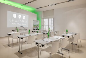 Una sala conferenze con tavoli e sedie bianche e uno schermo di proiezione che mostra il nome e il logo dell'hotel.