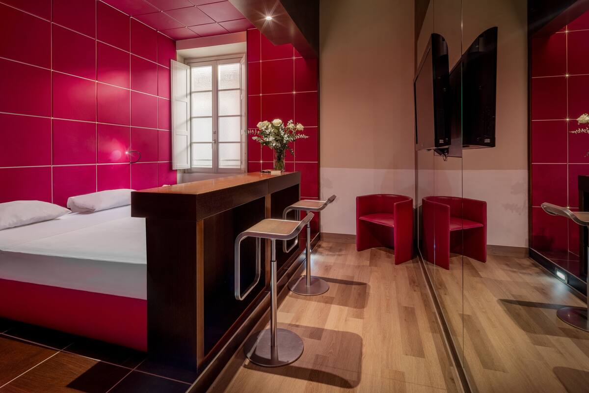 A modern hotel room with a large bed, a wooden desk with a vase of flowers, red chairs, and a large mirror.