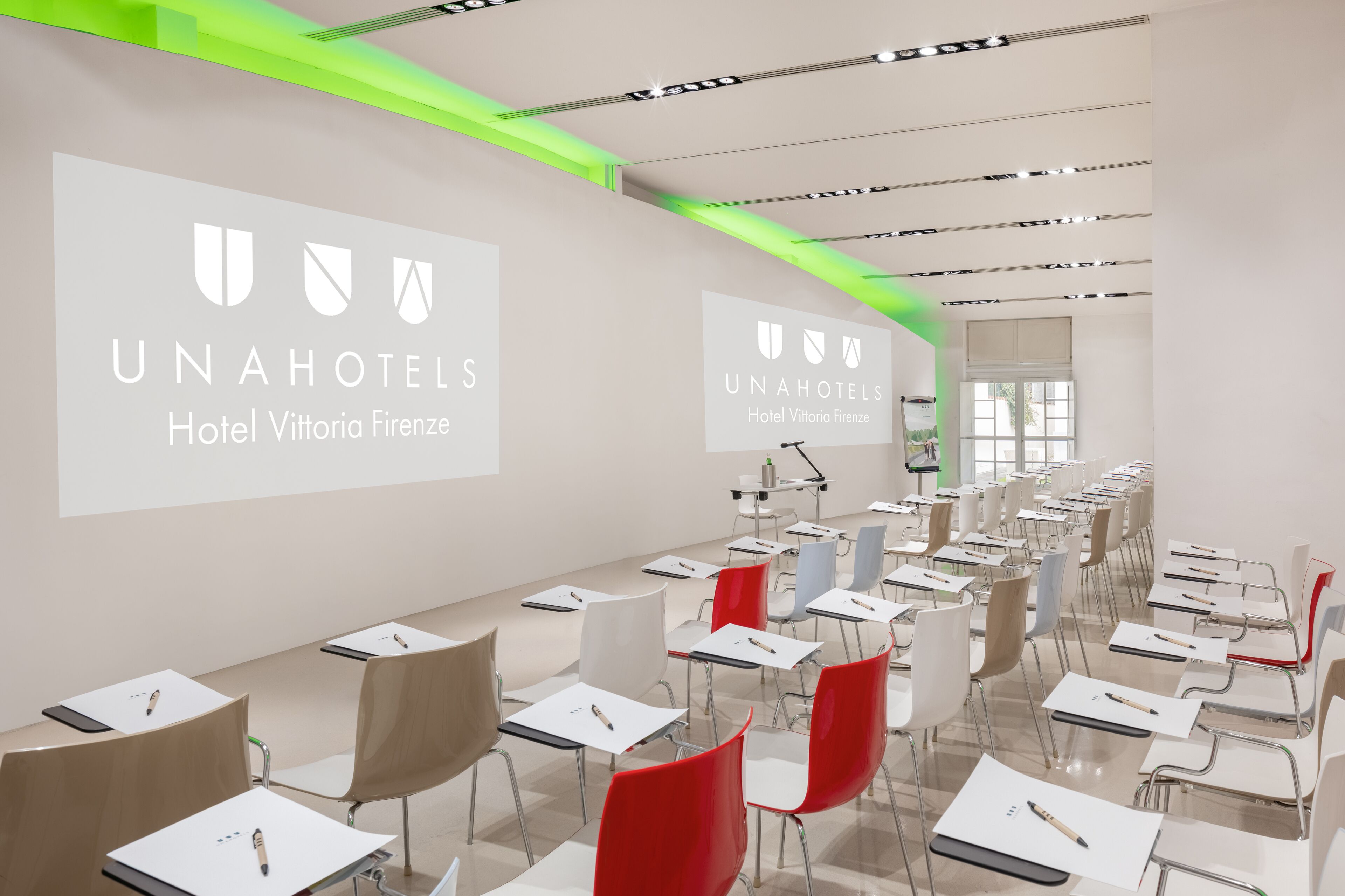 A conference room with rows of tables and chairs, a projector screen displaying the hotel name, and a bright green accent on the ceiling.