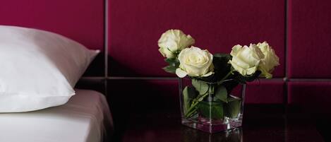 A bed with white bedding, a white pillow, and a glass vase with white flowers on a wooden nightstand against a tiled wall.