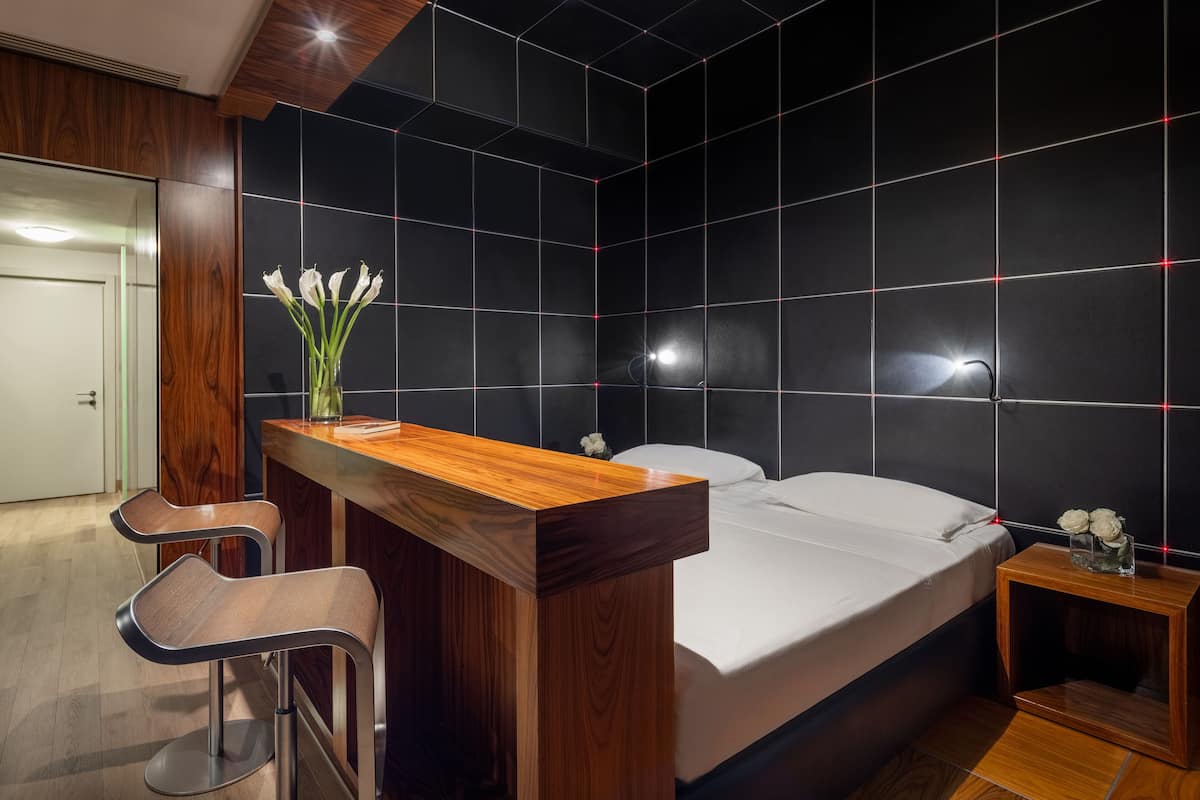 A modern hotel room with a wooden desk, two bar stools, a bed, and a nightstand with a vase of flowers.