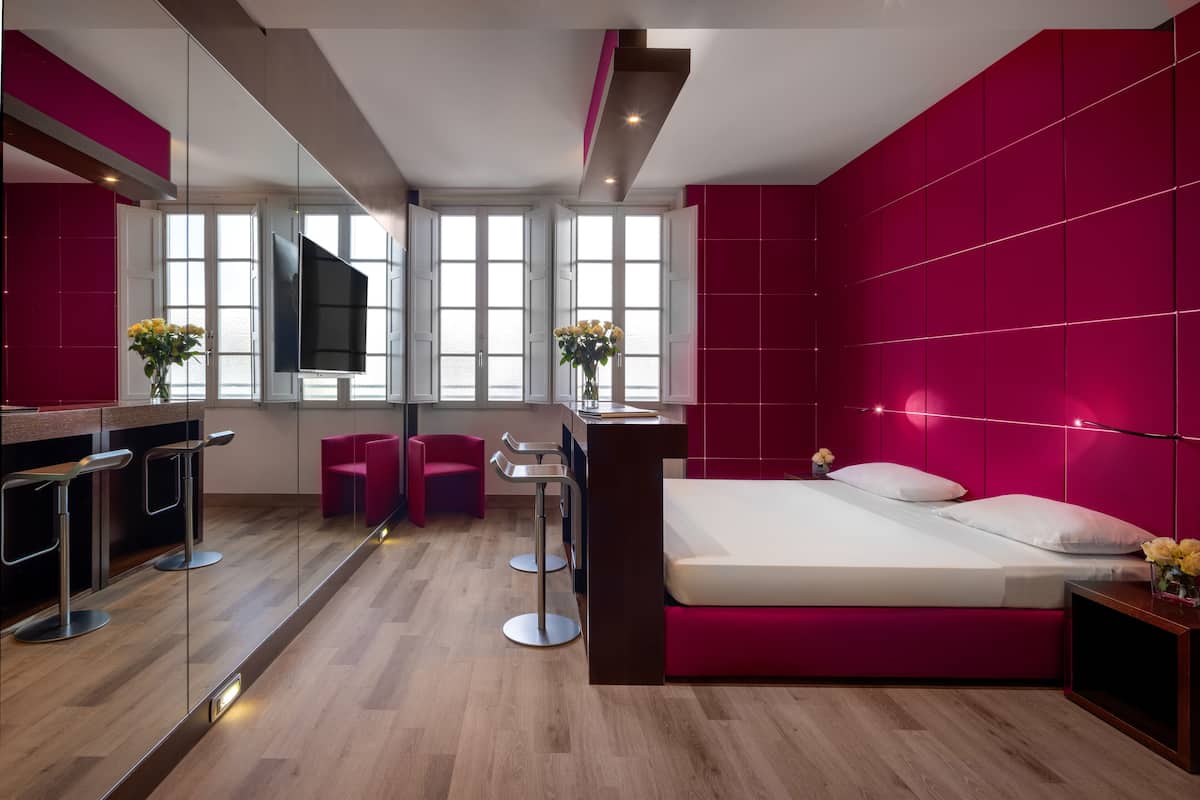 A modern hotel room with a large bed, a TV, a desk with a chair, and a bar area with stools.