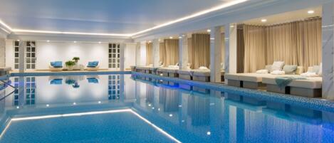 An indoor swimming pool with clear blue water, surrounded by lounge areas with seating and curtains.