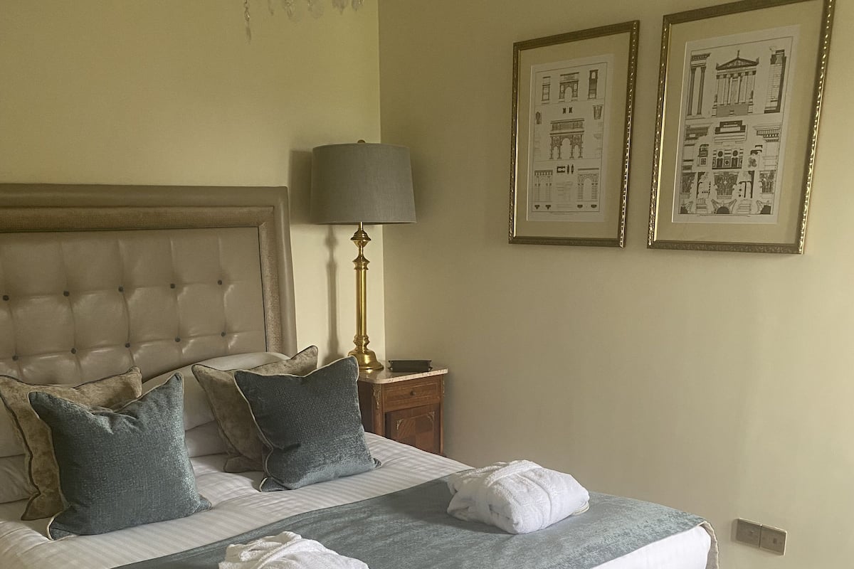 A neatly made bed with pillows, a bedside lamp, and a bench. There are two framed pictures on the wall.