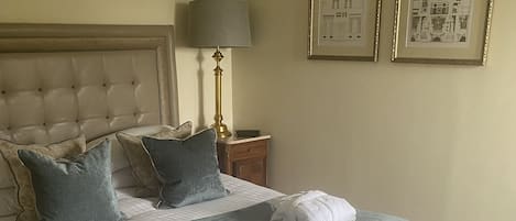 Una cama con sábanas bien puestas, almohadas, una lámpara de noche y un banco. Hay dos cuadros en la pared.