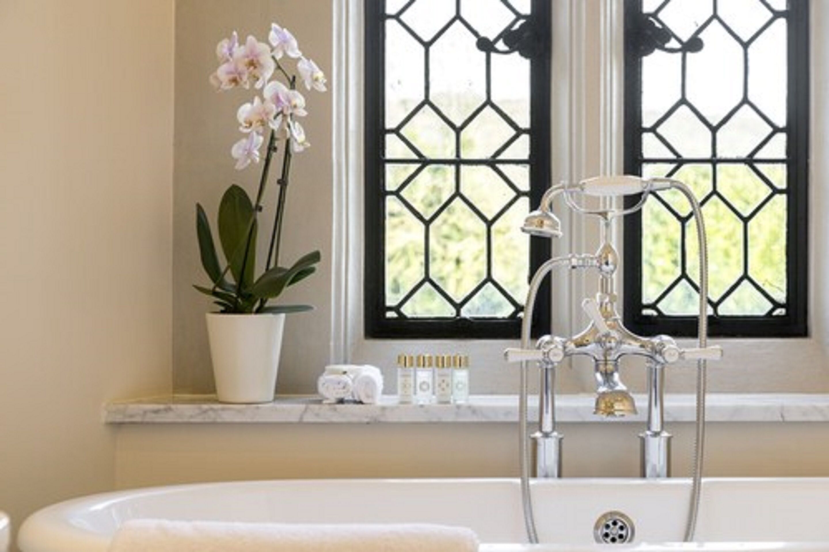 A bathroom with a bathtub, a window with a geometric pattern, a potted orchid, and toiletries on the windowsill.