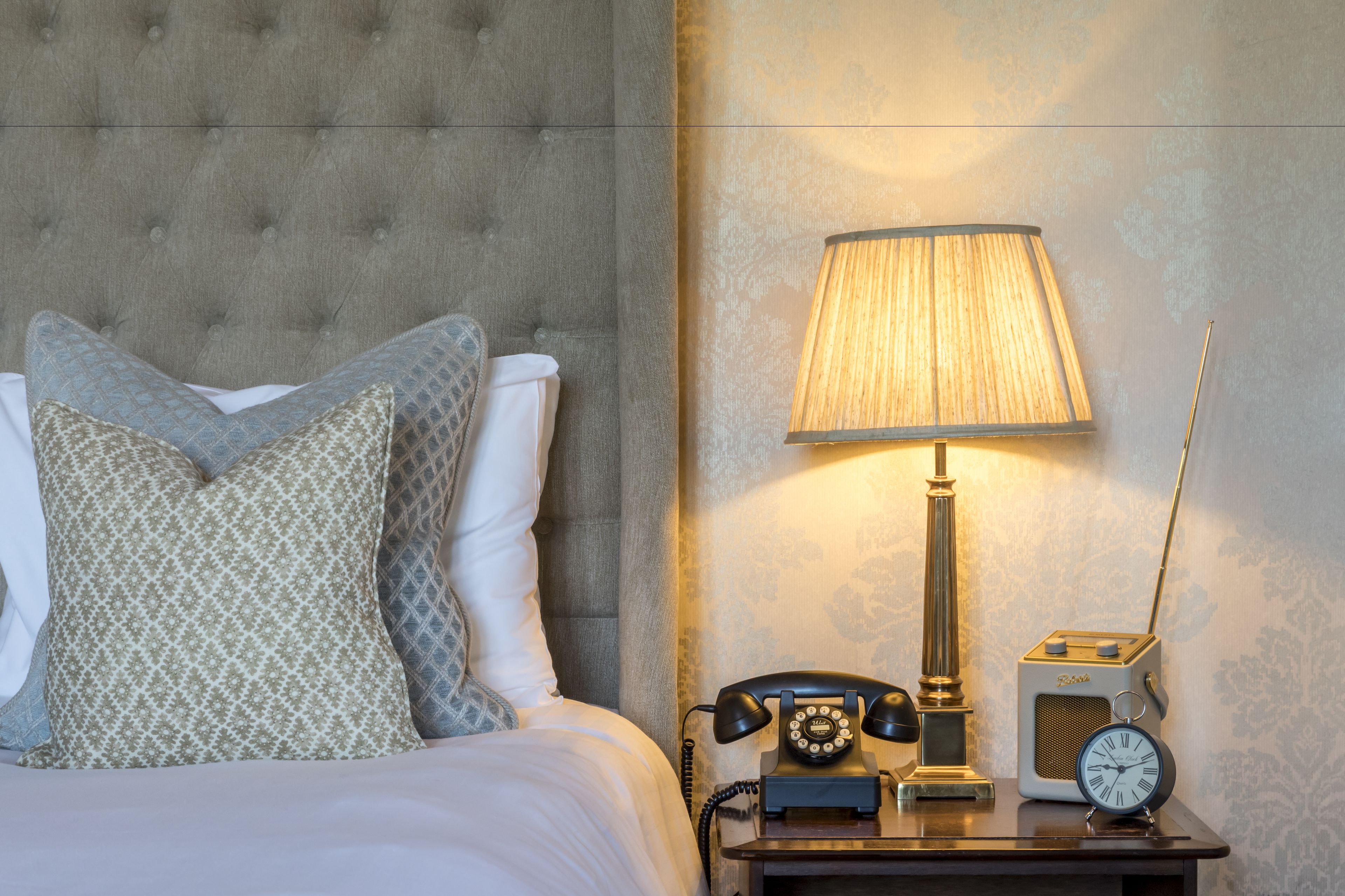 A neatly made bed with patterned pillows, a classic lamp, a vintage telephone, and a radio on a bedside table.