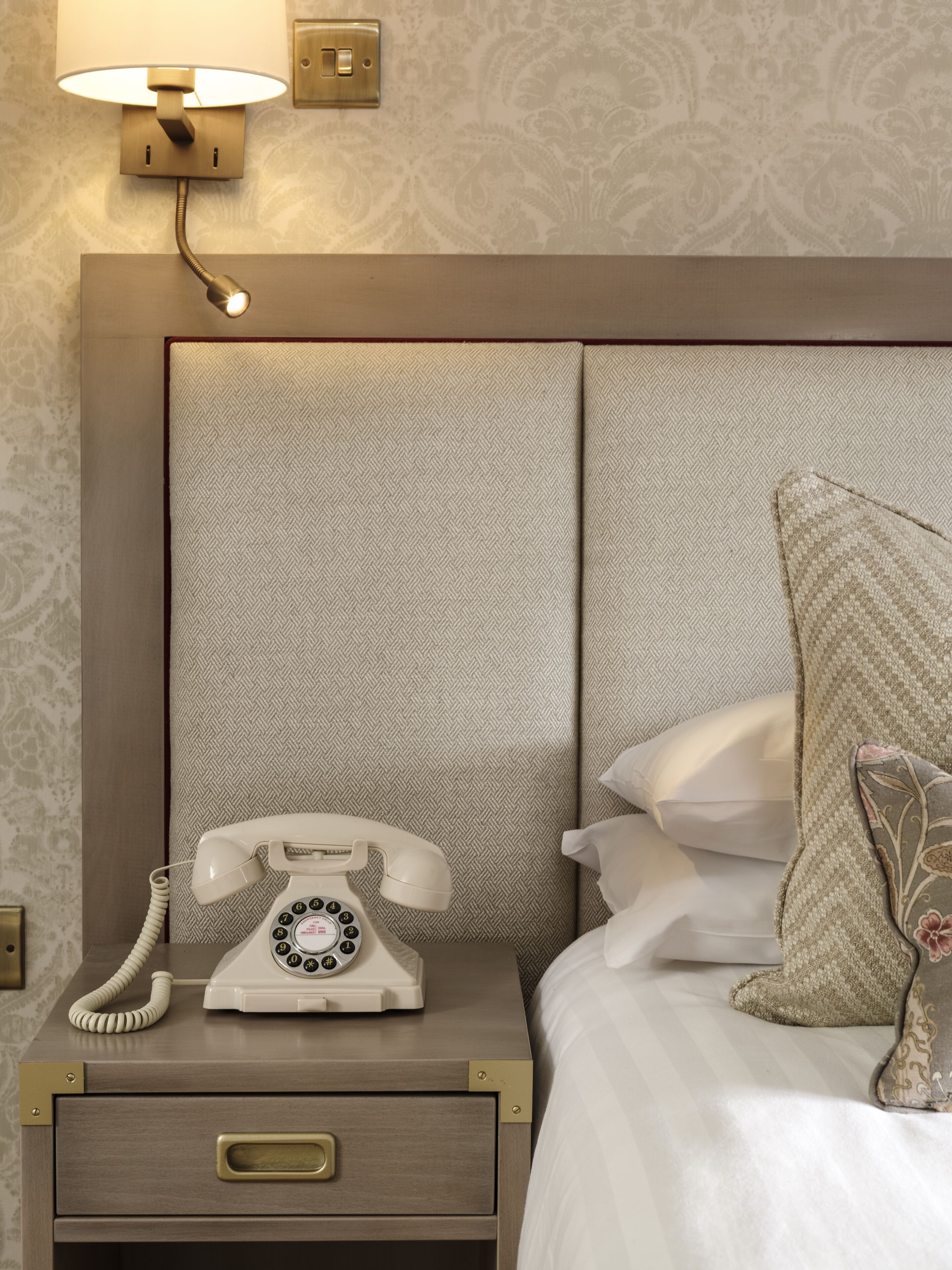 A hotel room with a bed, a bedside table with a rotary phone, a nightstand, and a wall-mounted lamp.