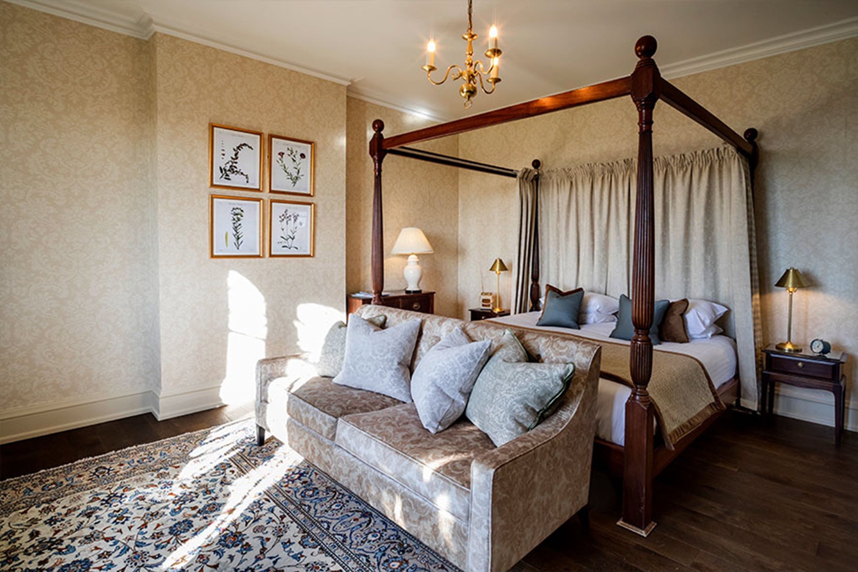 Een hemelbed met een baldakijn, een bank met kussens, een nachtkastje met een lamp, en ingelijste botanische prints aan de muur.