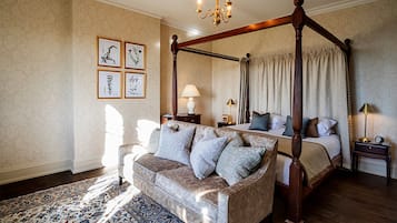 Een hemelbed met een baldakijn, een bank met kussens, een nachtkastje met een lamp, en ingelijste botanische prints aan de muur.