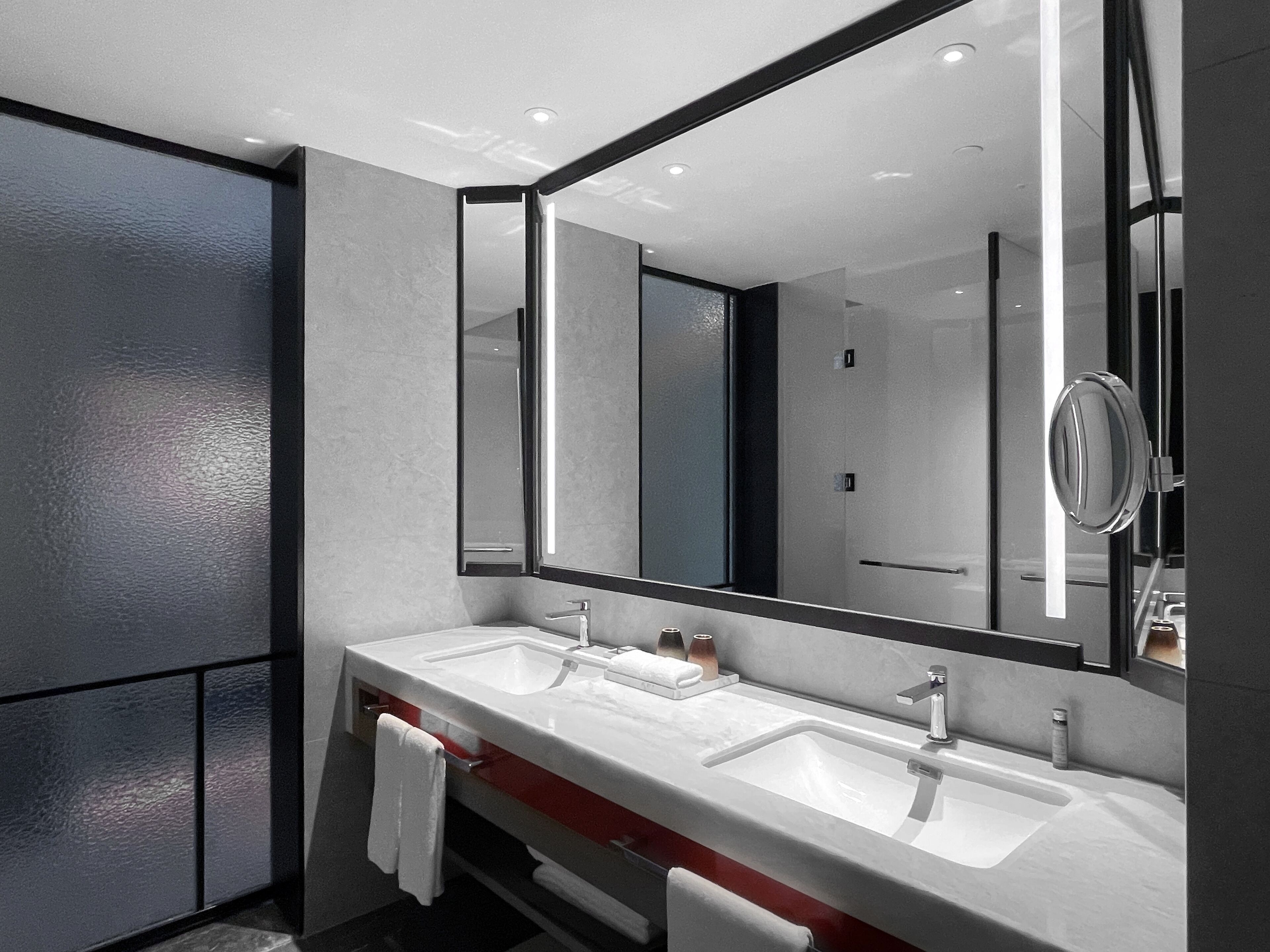 A modern bathroom with two sinks, a large mirror, and a red accent on the vanity.
