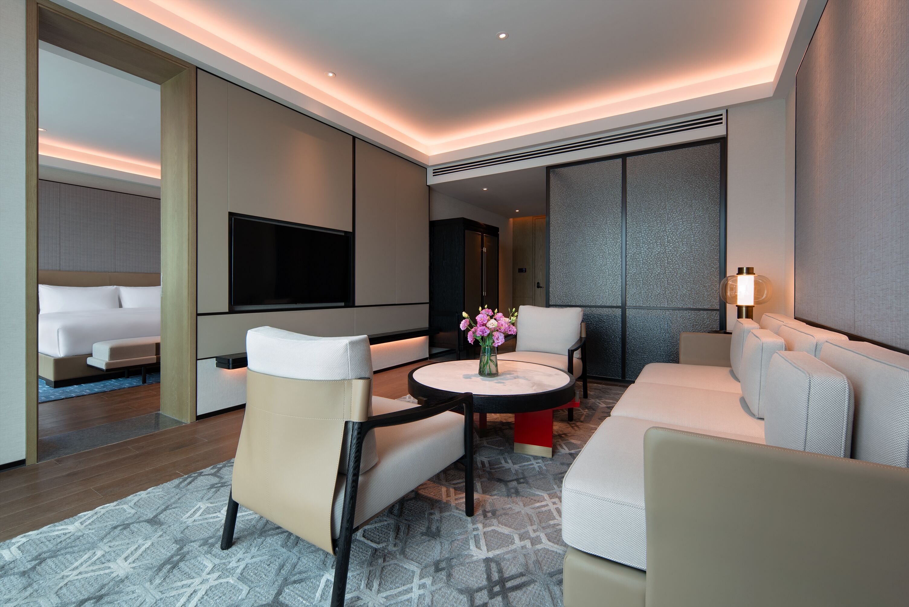 A modern hotel room with a bed, a TV, a seating area with a table and chairs, and a decorative vase with flowers.