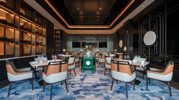 Un restaurante moderno con una mesa verde central rodeada de sillas anaranjadas y blancas, una alfombra azul estampada y una decoración oscura de estilo industrial.