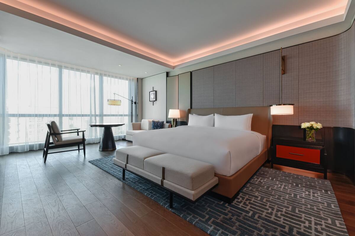 A modern hotel room with a large bed, a desk, a chair, and a view of the city.