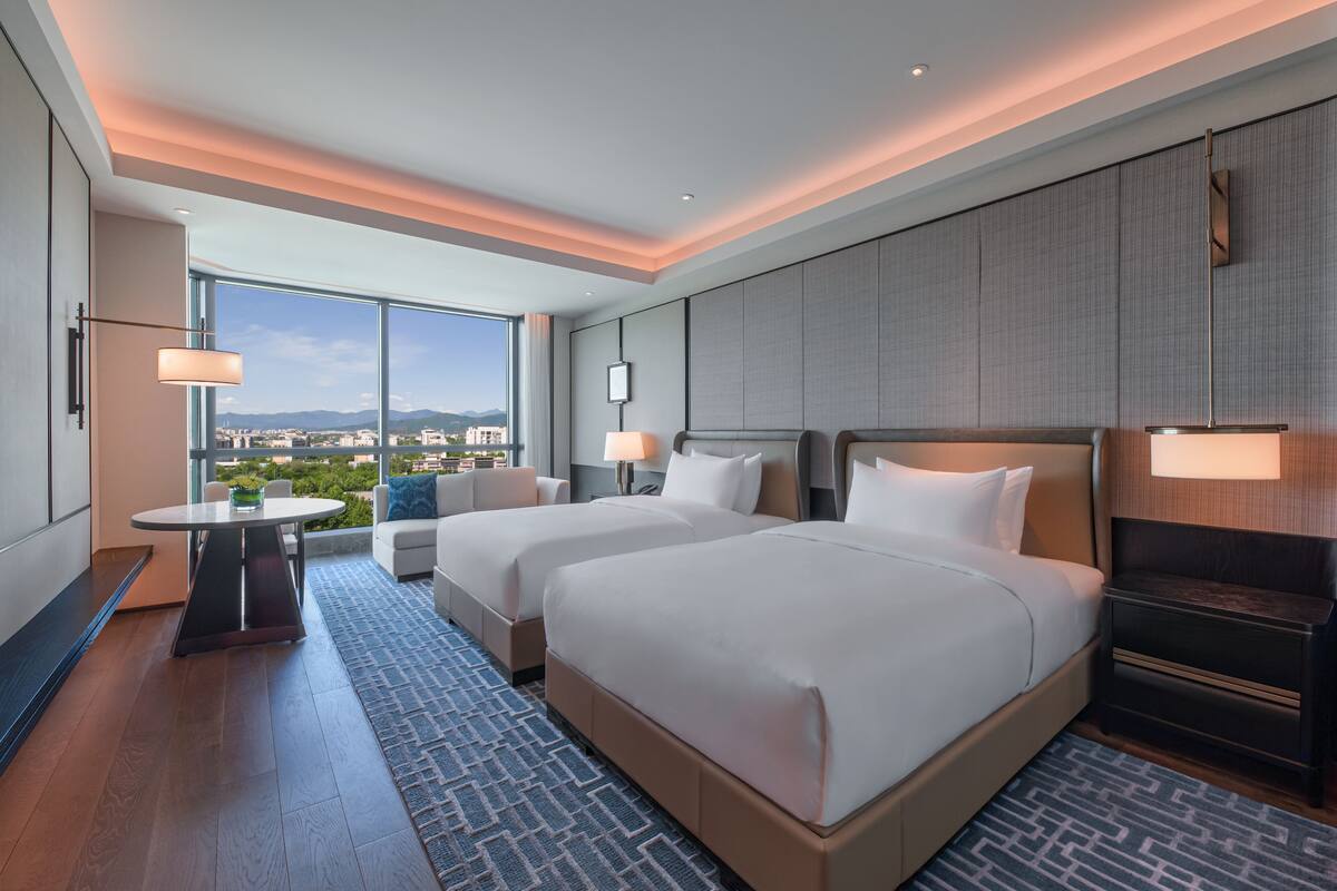 A hotel room with two beds, a small table, and a view of the city.
