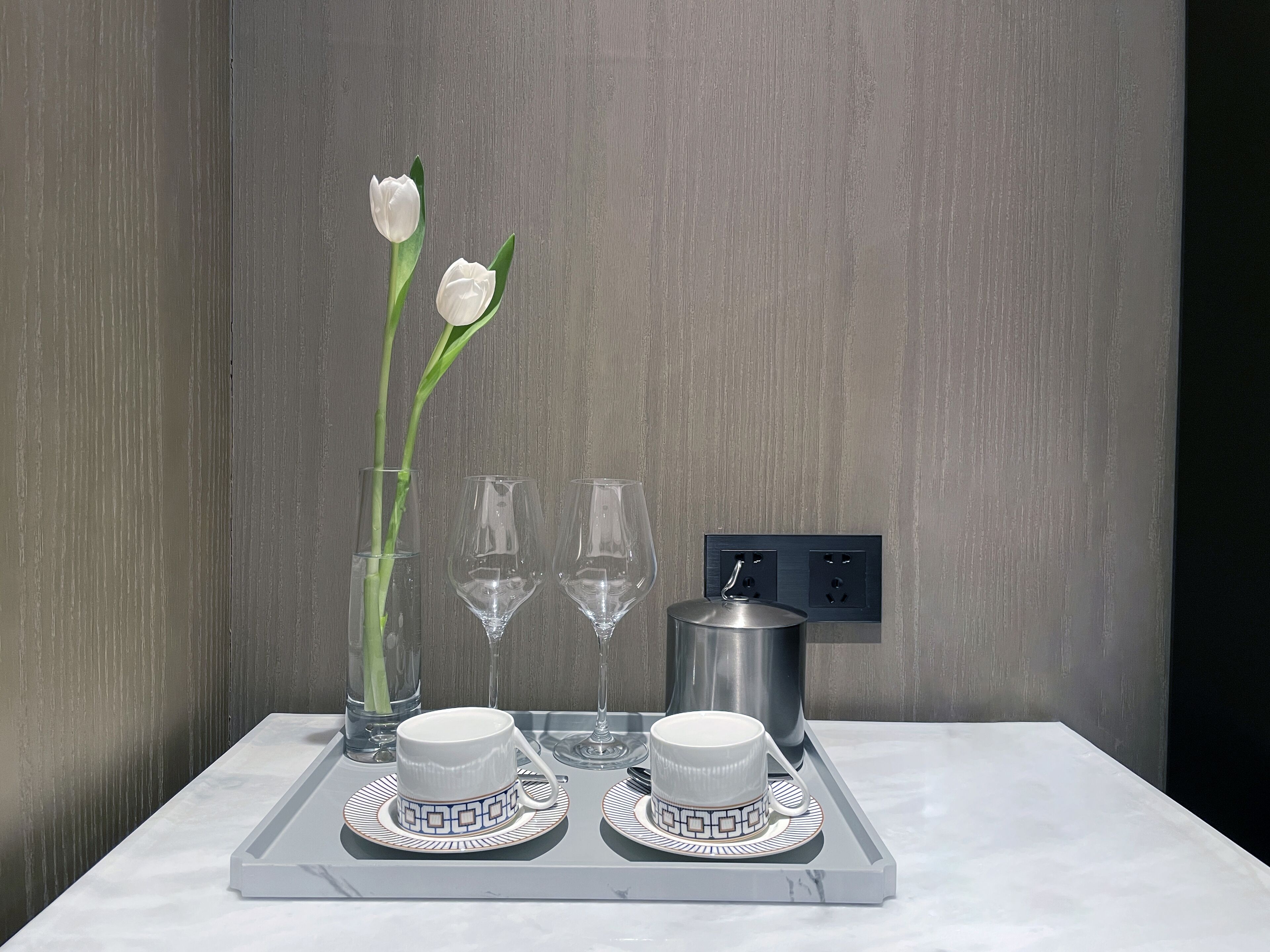 A tray with two white cups and saucers, a vase with white flowers, and three empty wine glasses on a marble surface.