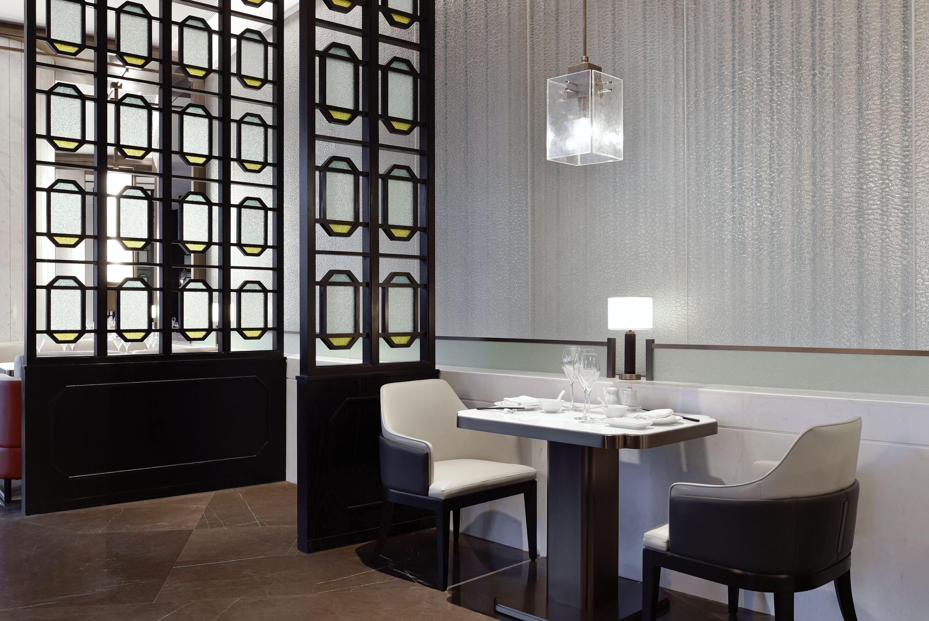 A modern dining area with a table set for two, a decorative partition with geometric patterns, and a hanging light fixture.