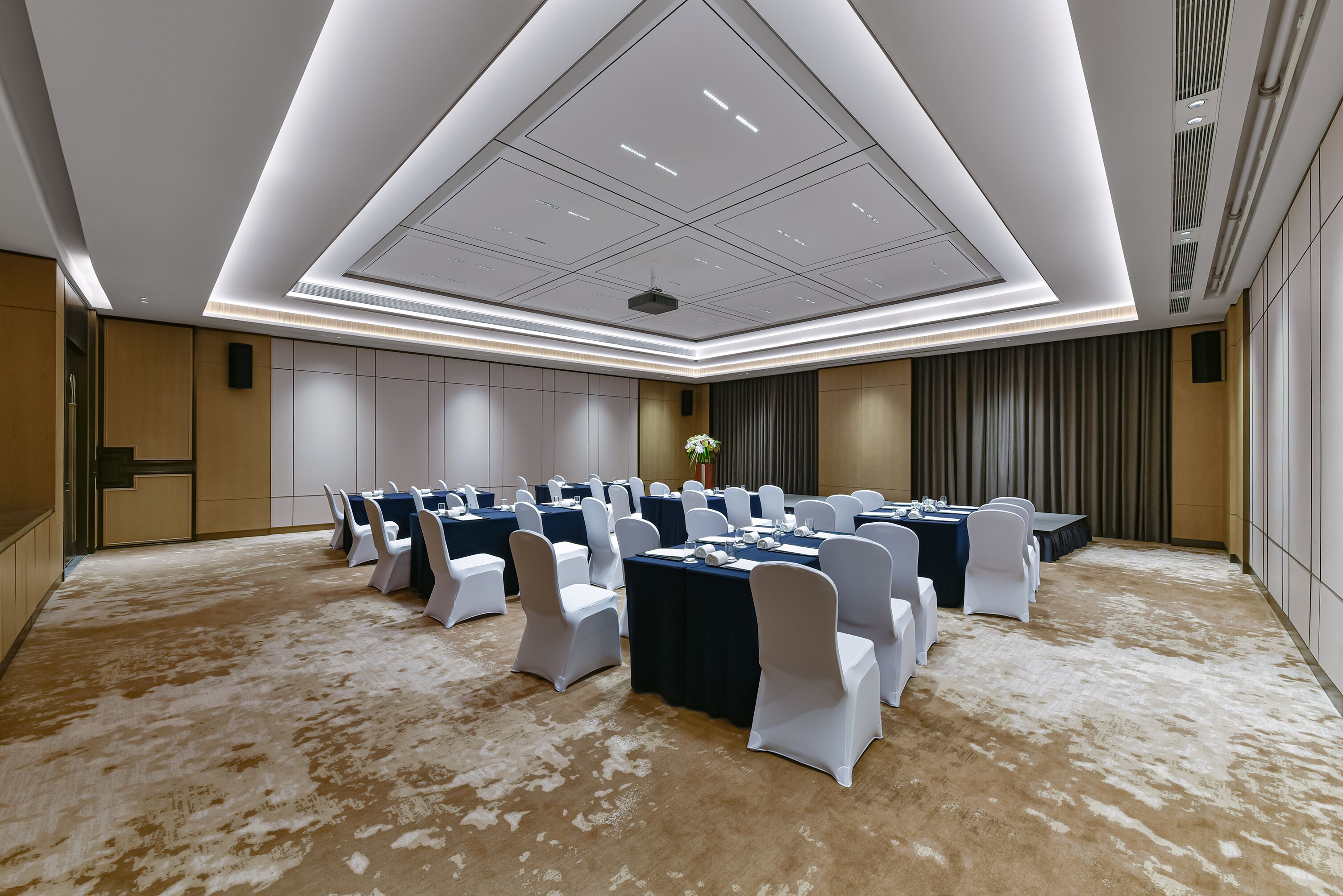 Un salón de conferencias con varias mesas y sillas, piso alfombrado y un diseño moderno en el techo.