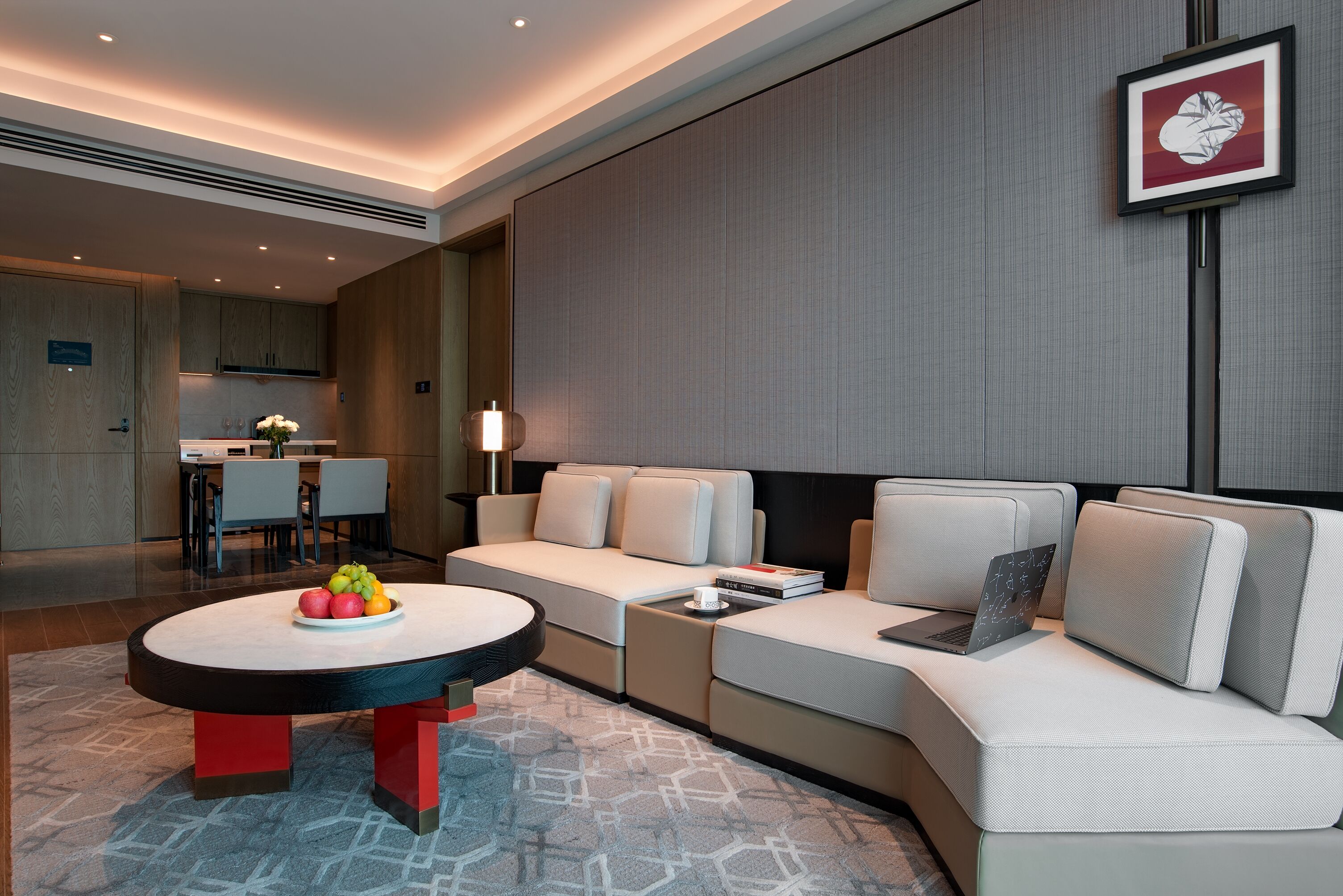 A modern hotel room with a sofa, a round table with fruit, a laptop, and a framed picture on the wall.