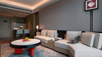 A modern hotel room with a sofa, a round table with fruit, a laptop, and a framed picture on the wall.