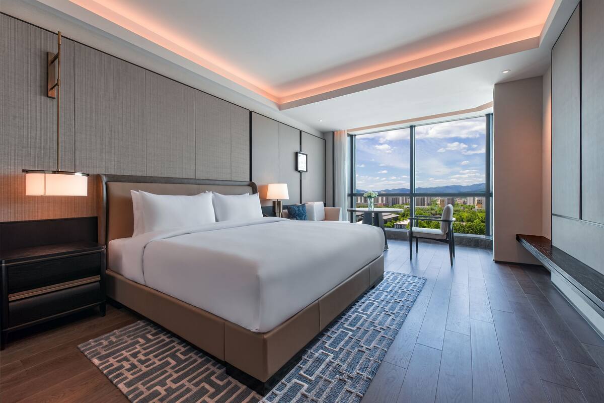 A modern hotel room with a large bed, a nightstand, a desk with a chair, and a balcony with a city view.
