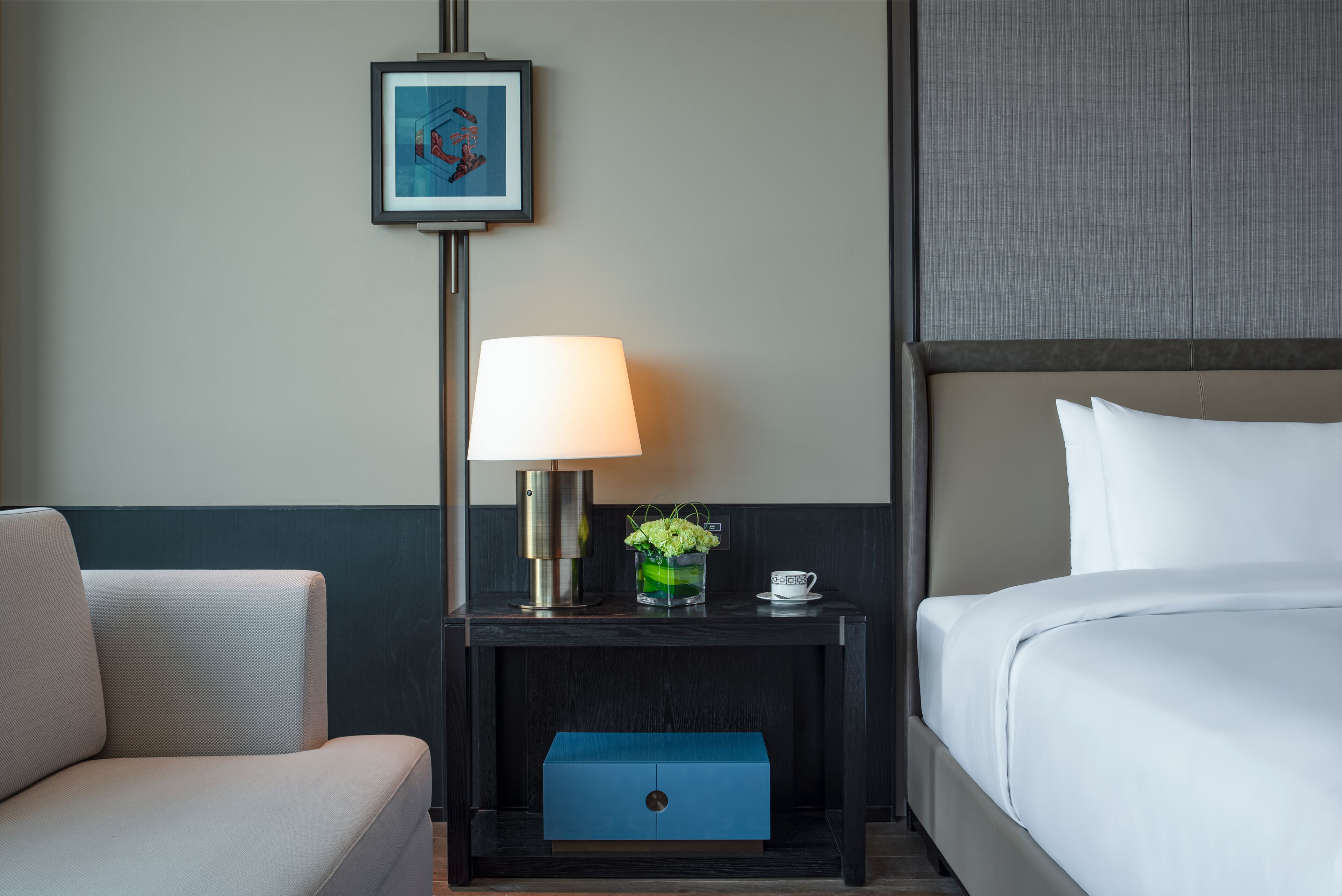 A hotel room with a bed, a nightstand with a lamp, a framed picture, a vase with flowers, and a small blue box.