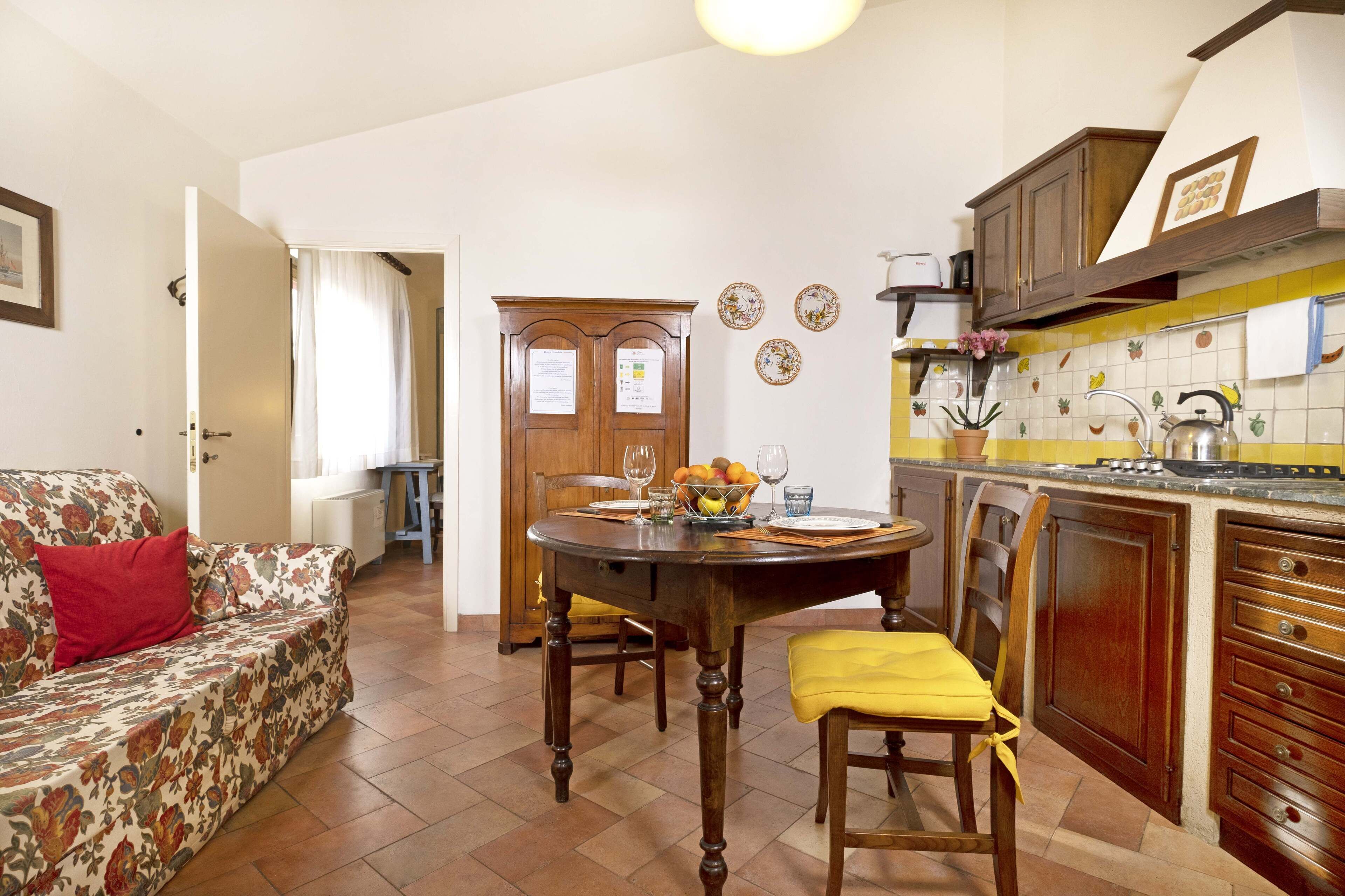 A cozy living room with a floral sofa, a dining table with chairs, and a kitchen area with wooden cabinets and a tiled backsplash.
