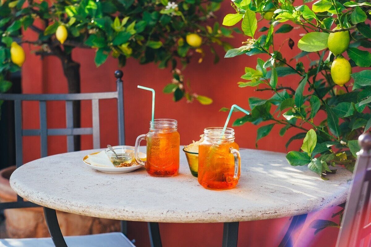A round table with two mason jars containing a beverage, a plate with food, and a bowl of fruit, set against a red wall with a lemon tree in the background.