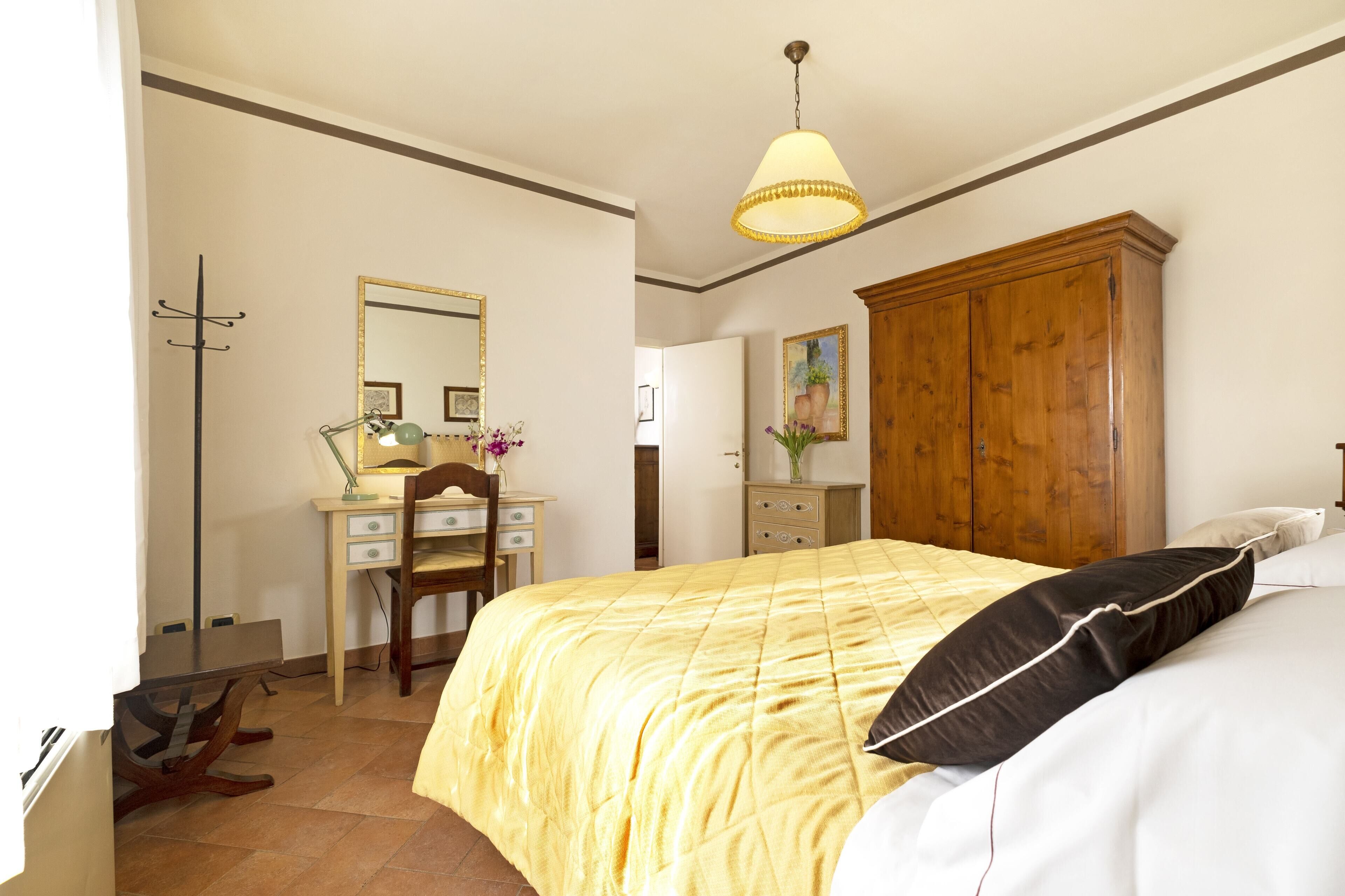 Une chambre à coucher comprenant un lit, une armoire en bois, un bureau avec une chaise, un miroir et une lampe suspendue.