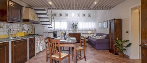 Een keuken met houten kasten, een eettafel met stoelen, een trap en een zithoek met een bank en een potplant.