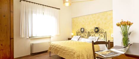 A bedroom with a bed, wooden wardrobe, a window with curtains, a bedside table with a lamp, and a vase with flowers.