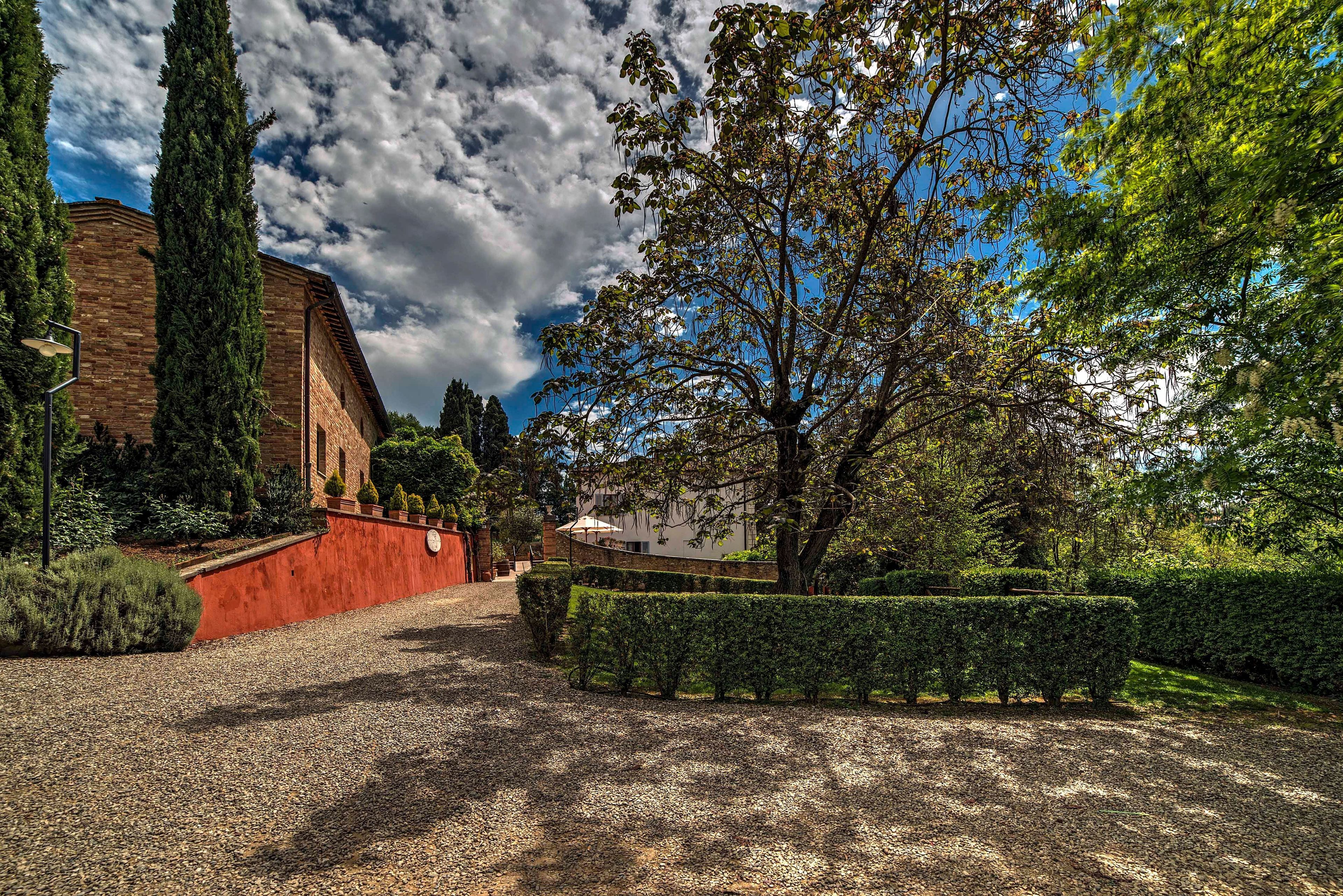 A gravel pathway leading to a building with a red wall and a large tree in the foreground.