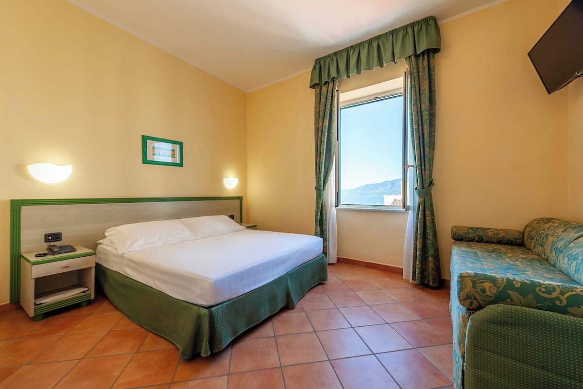 A hotel room with a bed, a nightstand with a phone, a small table with a lamp, a window with curtains, and a sofa.