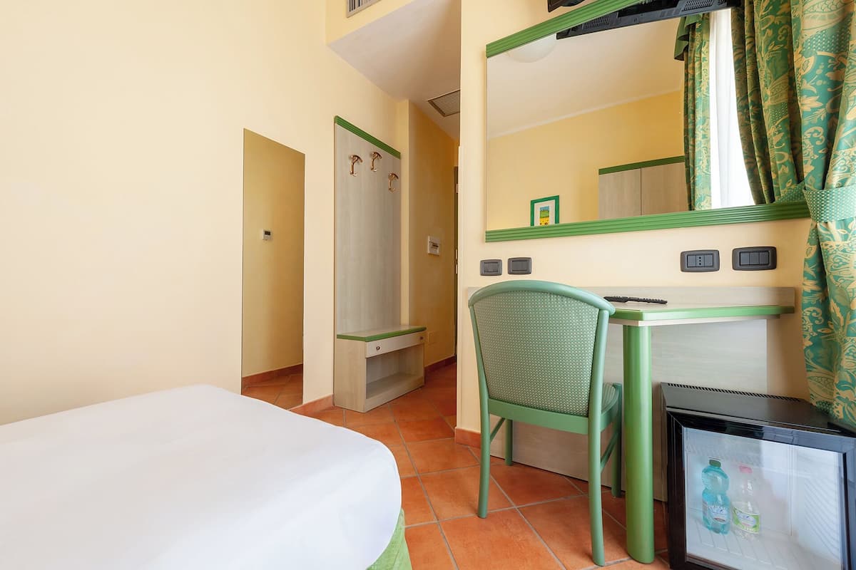 A hotel room with a bed, a desk with a chair, a mirror, and a refrigerator.