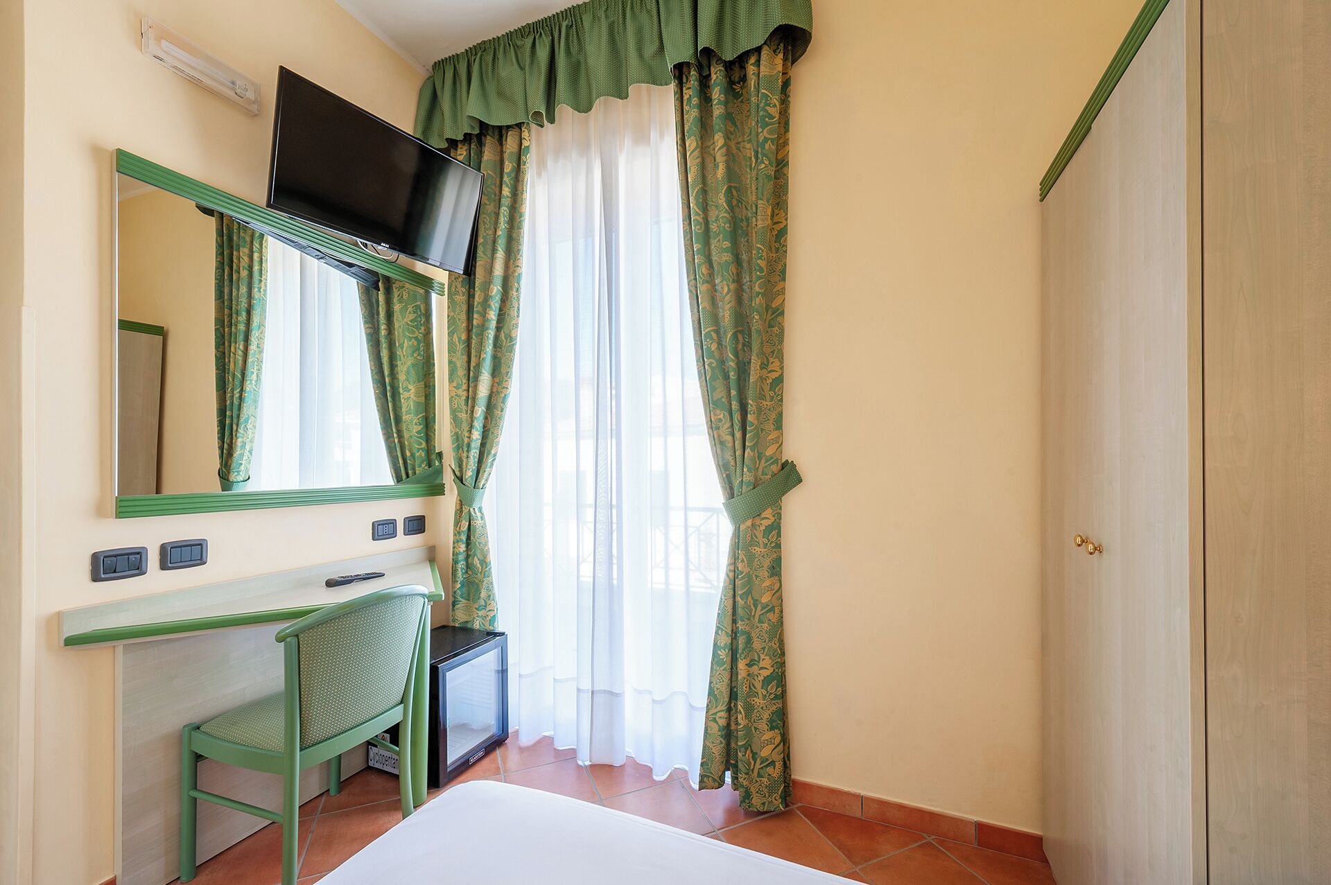 Une pièce comprenant un lit, un bureau avec une chaise, un téléviseur fixé au mur et une porte.