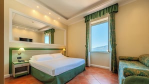 Habitación de hotel con cama, mesita de noche con lámpara, sofá, ventana con cortinas y espejo en la pared.