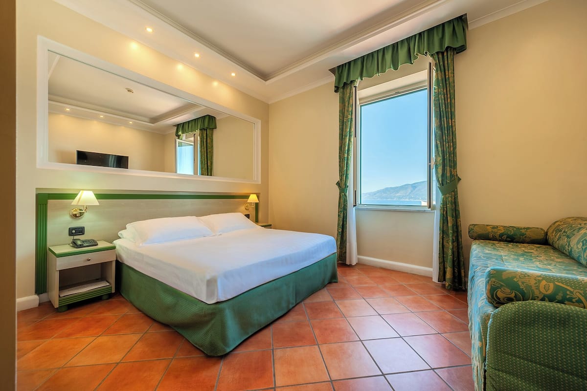 A hotel room with a bed, a nightstand with a lamp, a sofa, a window with curtains, and a mirror on the wall.